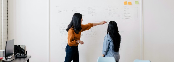 Two women with long hair standing in front of a whiteboard. One is wearing an orange shirt and pointing to something written on the board.