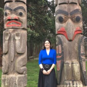 Jennifer Ward in a blue, long sleeved top and long black skirt, standing in between two totem poles.