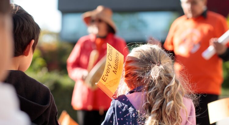 The back of a child’s head, whose hair is pulled up into a ponytail. They are holding a flag that reads “Every Child Matters” and is watching two people wearing orange in a ceremony for Truth and Reconciliation day at Langara College.