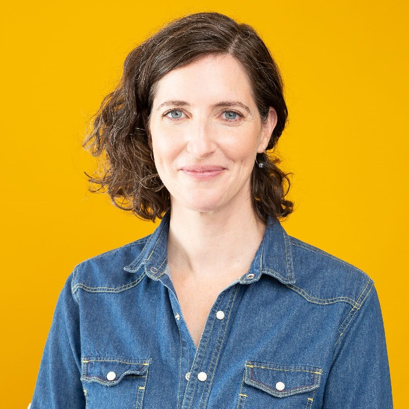 Headshot of Alex Samur, who has brown hair and is wearing a denim top. The background is a bright yellow-orange.