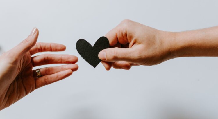 Close-up of one hand reaching for a small paper heart that is being offered by another hand.