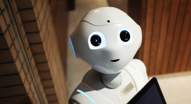 White robot with large round eyes and a small lined mouth looking up at the camera. The robot has some areas of blue light showing through near its ear and shoulder, and appears to have a screen attached to its chest.
